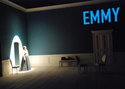 woman standing in spotlight, text projected onto wall that reads "Emmy"