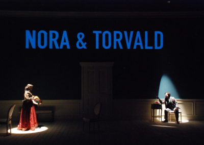 two people in spotlights, text projected onto wall that reads "Nora & Torvald"