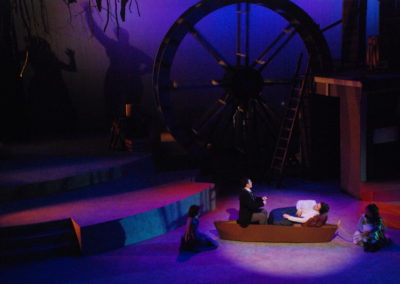 two people in boat, two others sitting next to the boat on the floor, stage view of mill wheel and shadows on backdrop