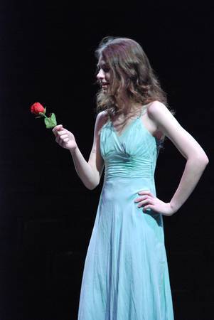 a woman stands in a spotlight holding a rose