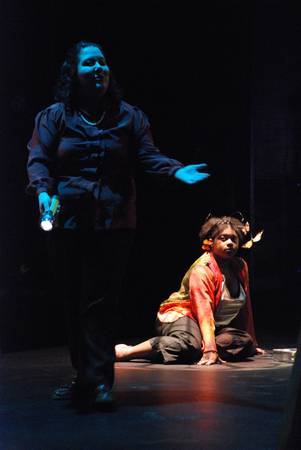 a shadowed woman standing in the foreground, a woman sitting on the floor in a spotlight behind the first woman