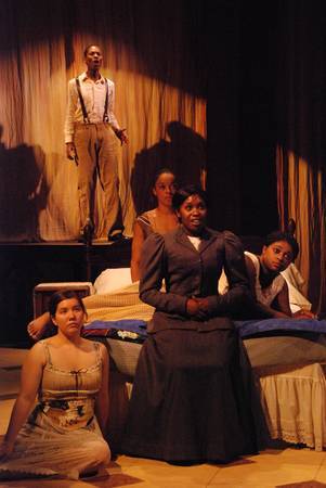 a man in a spotlight on a platform upstage, a woman sitting on the edge of a bed with three other women around her