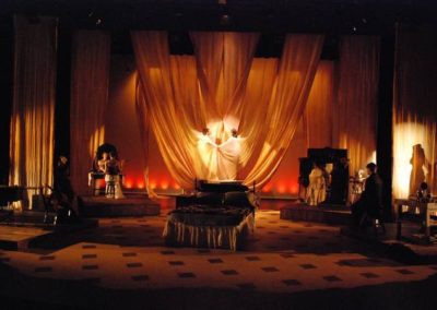 women creating sculpture out of hanging fabric center, silhouette of a woman stage left, three other people sitting in various places on stage in the shadows