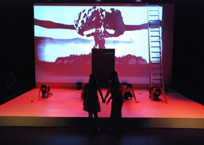 silhouette of two people in the foreground, 4 women crawling on hands and knees, projected image of mushroom cloud on backdrop