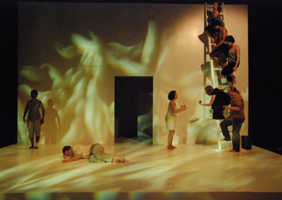 five people climb a ladder on stage left, a woman stands near the ladder talking to them, a man lays on the floor center, a woman stands in the shadows stage left