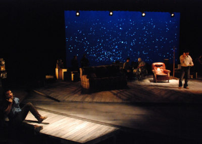 two men talking on telephones, one at a payphone stage right and the other on a house phone stage left, a woman in a wheelchair is upstage with another man sitting next to her