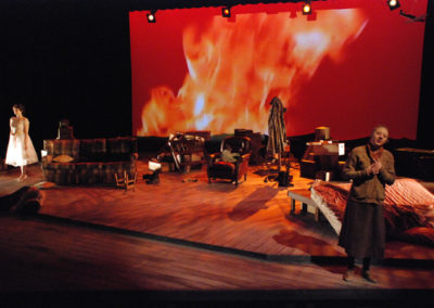 two women onstage separated by a good distance, woman stage right is standing in a spotlight, woman stage left is talking to the audience, projected image of fire on the backdrop