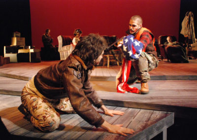 a man on his hands and knees in the foreground, another man holding the American flag crouches and points a shotgun at the first man, three others sit in the background unaware of what's happening