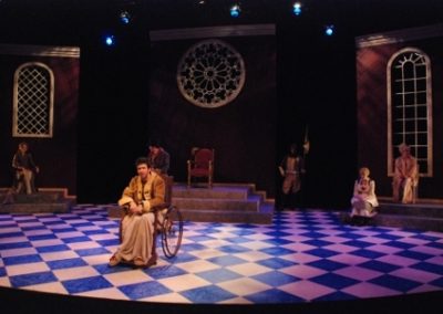 the king in a wheelchair addressing the audience, his court of five people sit upstage in the shadows listening