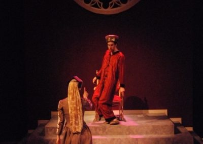 a woman gesturing towards the king who is up on a platform near his throne