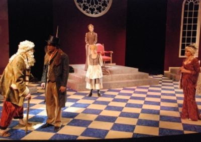 two men stand talking on stage right, three women watch from the side