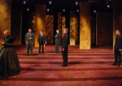 a woman stands downstage right, five men stand in various places upstage and around all looking at her