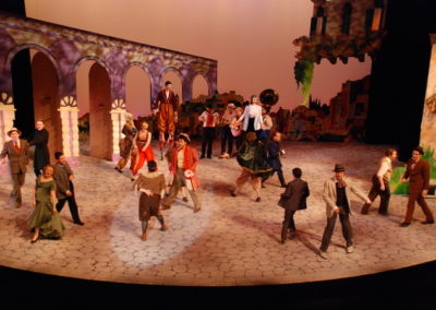 full stage activity, twenty people on stage in various small groups doing various things, including a small strolling band, a man on stilts, and a juggler
