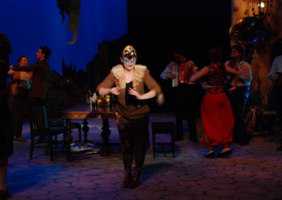 a woman wearing a Venetian mask stands in a spotlight addressing the audience, several partygoers can be seen in the background