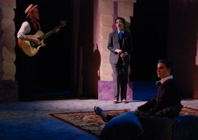 a man sits on the floor listening to music, a man stands playing guitar while a woman stands nearby singing