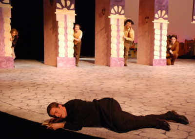 a man lies on the ground in despair, four people look on from upstage, all peaking out from behind the scenery