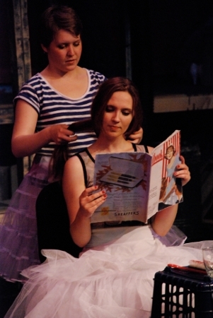 a woman sitting and reading a magazine, a woman standing and doing the other woman's hair
