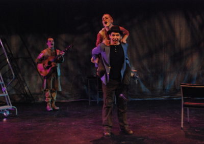 a man playing guitar upstage right, a woman riding piggyback on a man standing center stage
