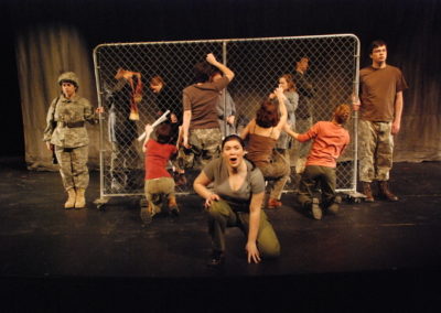 a clash of people at a chunk of chain-link fence, two soldiers each holding one of the sides of the fence, a woman crouched down center on one knee talking to the audience