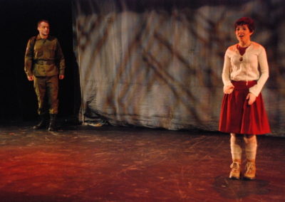 a woman stands alone stage left, a man looks on from stage right