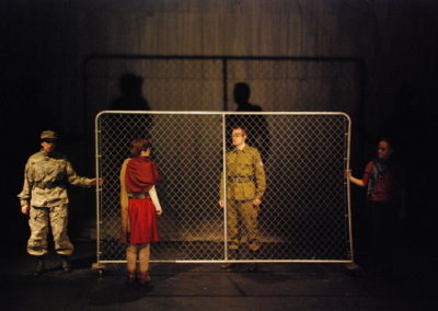 a man and a woman separated by a chain-link fence, two soldiers holding the fence on the edges