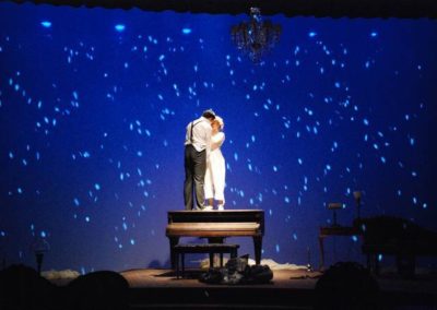 a man and a woman embrace in a spotlight standing on a grand piano, projected image of snow on backdrop all around them