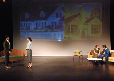 two scenes, a man and woman stand talking in front of a couch stage right, a man and a woman sit on a couch stage left, projected images of two different houses on backdrop