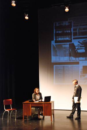 a man approaches a woman who is sitting at a desk, projected images of office items on the backdrop