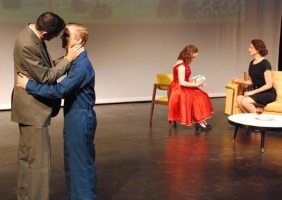 two men about to kiss, standing and talking downstage right, while two women sit and talk upstage left
