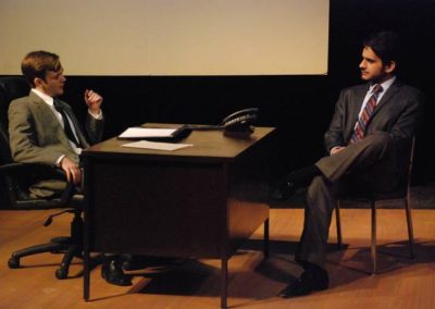 two men sitting across from one another, with a desk between them, talking