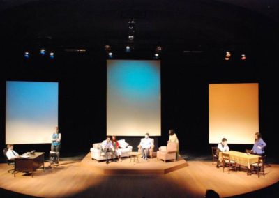 full stage showing three areas, two people in an office space stage right, four people in a living room center, two people in a dining room stage left