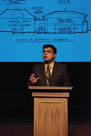 man standing at podium, projected image of an architectural blueprint on the backdrop behind him