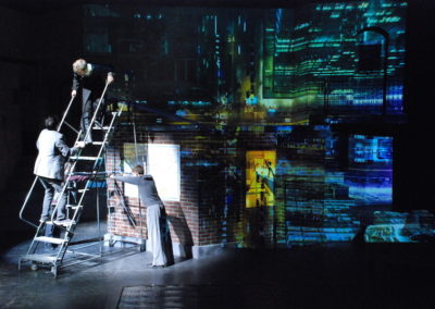 two people climbing a rolling stair unit to a taller platfoorm, image of cityscape projected across stage