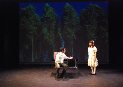 man sitting in chair gesturing to woman standing nearby, projected image of trees in a park on backdrop