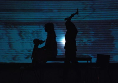 silhouette of a man with an axe raised over his head standing behind a woman sitting on the edge of a table, projected image of glitched video on background