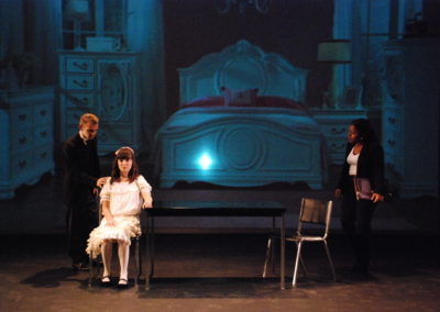 woman sitting at a table, man standing with his hand on her shoulder, other woman standing and watching, projected image of a girls bedroom on backdrop