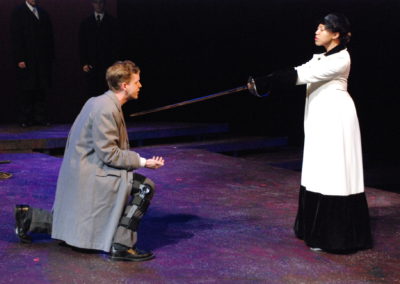 woman standing and holding a sword in her outstretched arms, while Richard kneels in front of her with his chest inches away from the tip of the blade