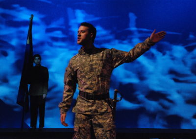 man standing in camouflage military fatigues gesturing with left hand, woman standing with flag watching