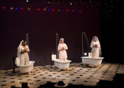 three women dressed as brides, each standing in a vintage bathtub, with showers running