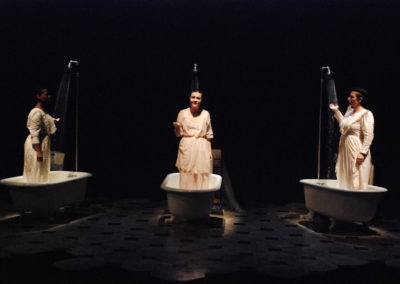 three women, each standing in a vintage bathtub, with showers running, outside two looking towards center