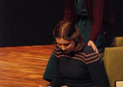 woman sitting on the ground looking at a yearbook, woman perched on the edge of a chair with her hands on the other woman's shoulders