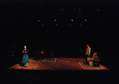 woman in spotlight stage right, man in shadows watching from stage left