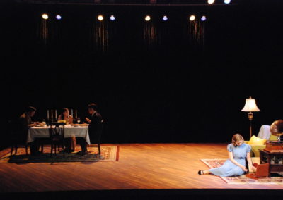 three people eating at the dinner table stage right, woman sitting alone stage left