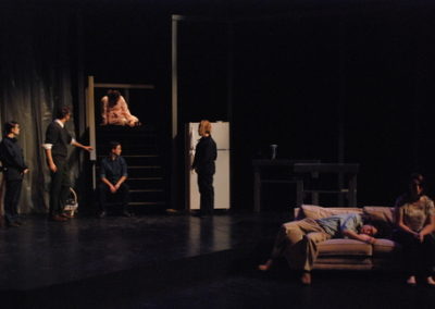 woman sitting on stair landing with 4 people around her upstage right, two people sitting on couch downstage left.