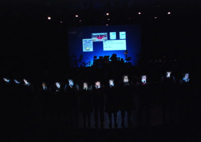 thirteen performers standing in the dark with faces lit by their cell phones