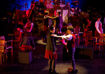 two people in a spotlight center about to dance, two more people upstage in a similar pose, others around the space watching